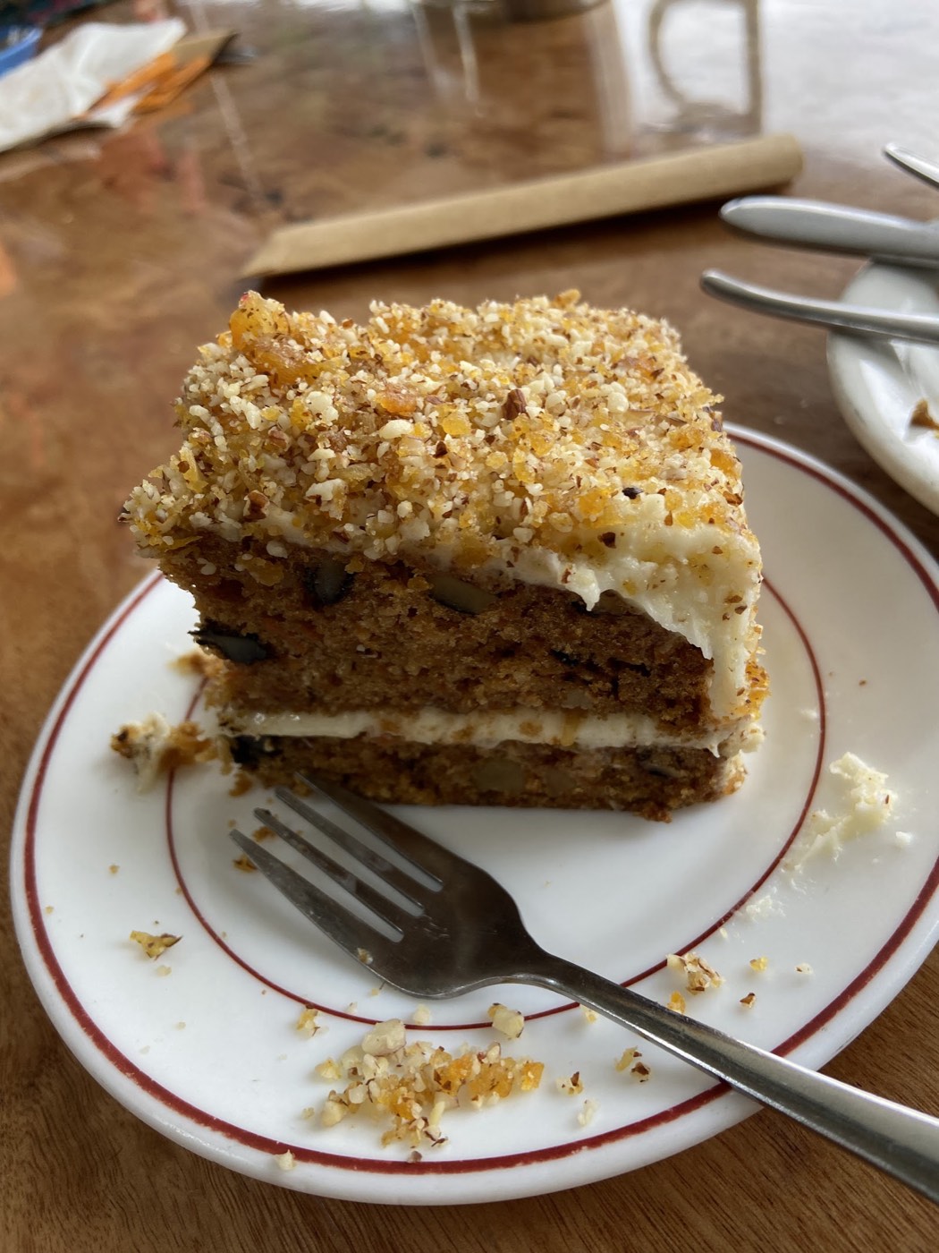 A slice of layered carrot cake with white icing between the layers and a crunchy nut topping, on a plate with a fork