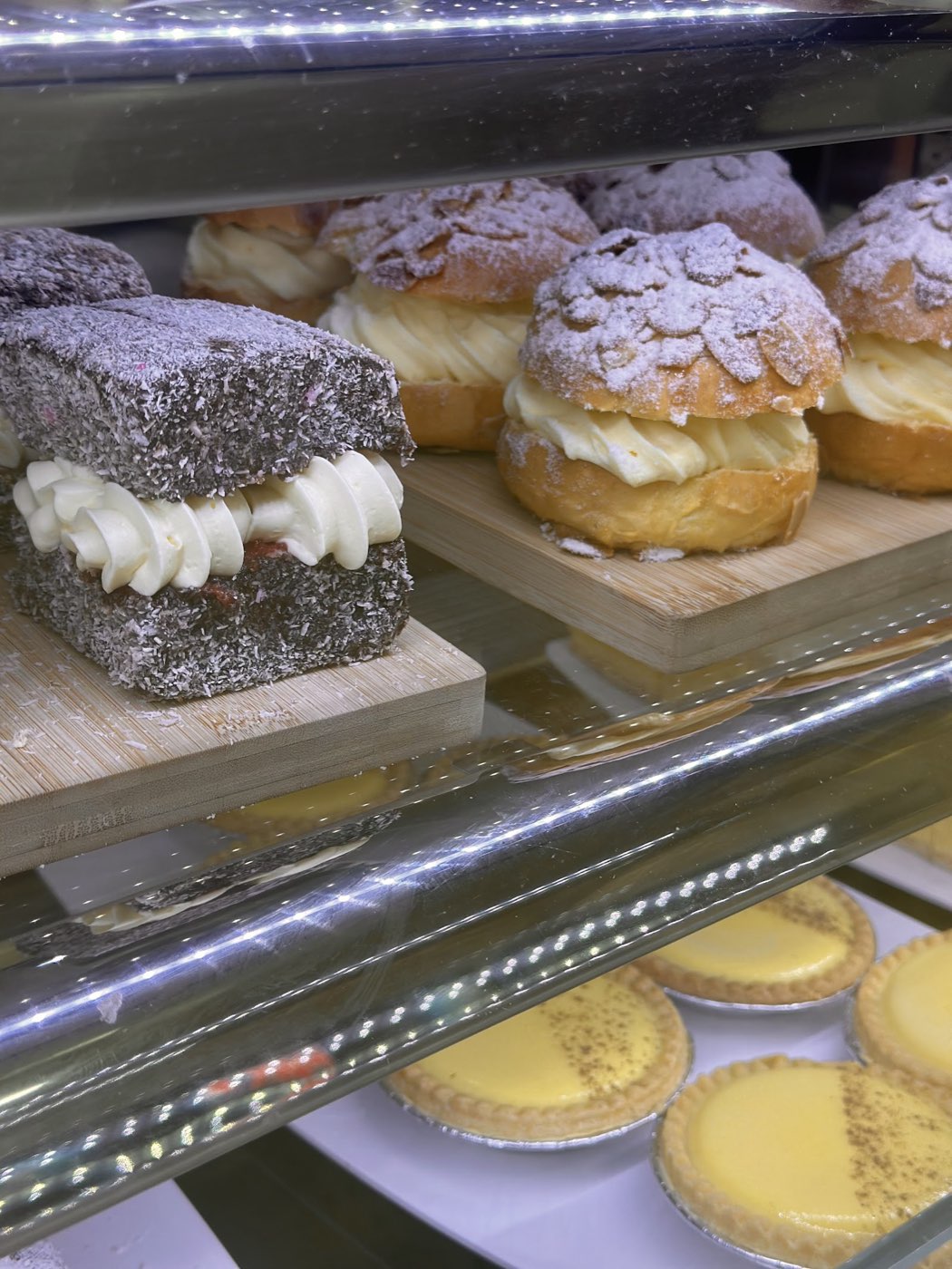 A cream-filled lamington beside sugar-dusted cream puffs in the cabinet, with yellow tarts on the shelf below